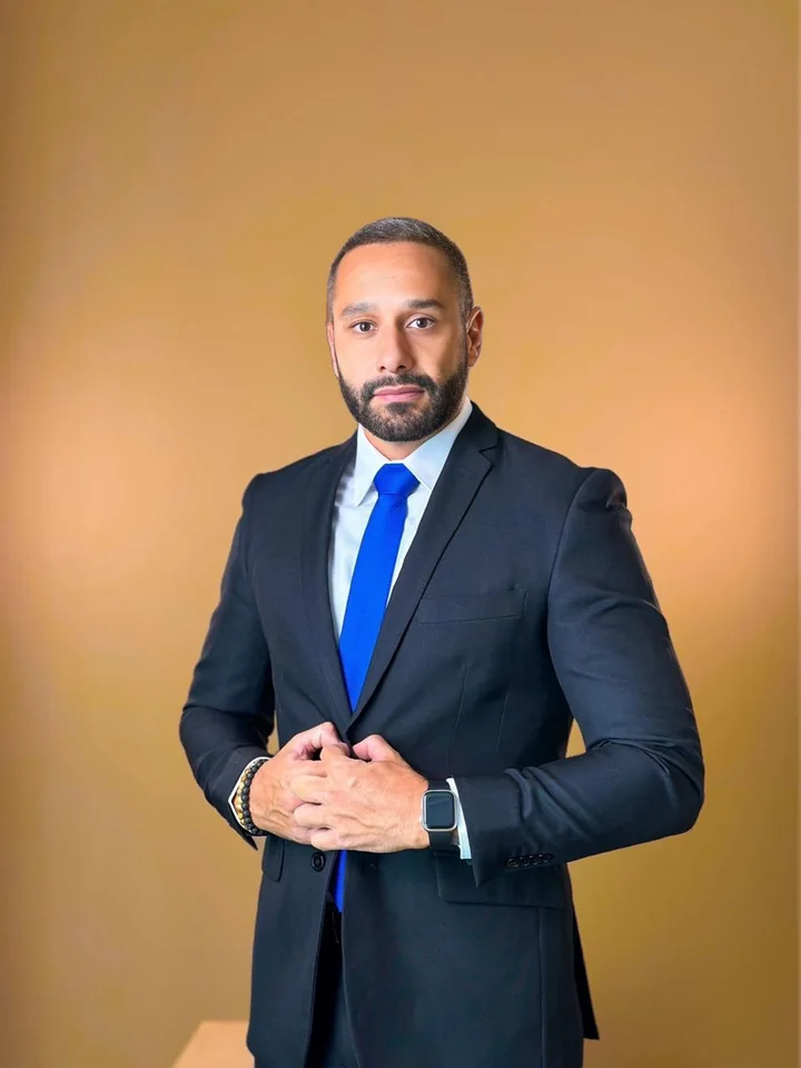 Dr. Arnaldo Candella de terno escuro e gravata azul, em frente a um fundo dourado